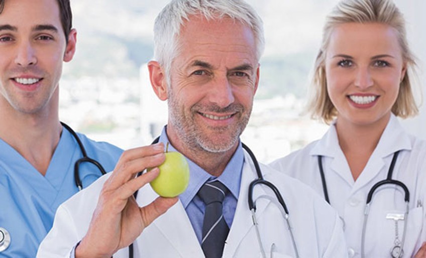 Krankenversicherung - zwei junge Ärzte stehen neben einem Oberarzt, der einen Apfel in der Hand hält.